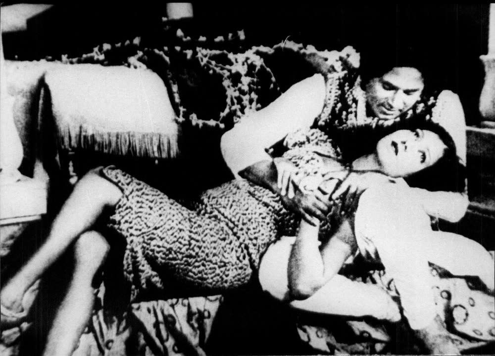 Black-and-white still from the 1931 film Alam Ara: a man leans over a woman reclining across a low couch, both in heavily patterned costume.