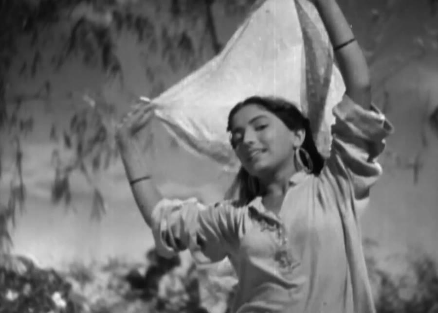 Black-and-white film frame: a young woman outdoors, smiling, holding her dupatta stretched taut above her head with both arms during a song.