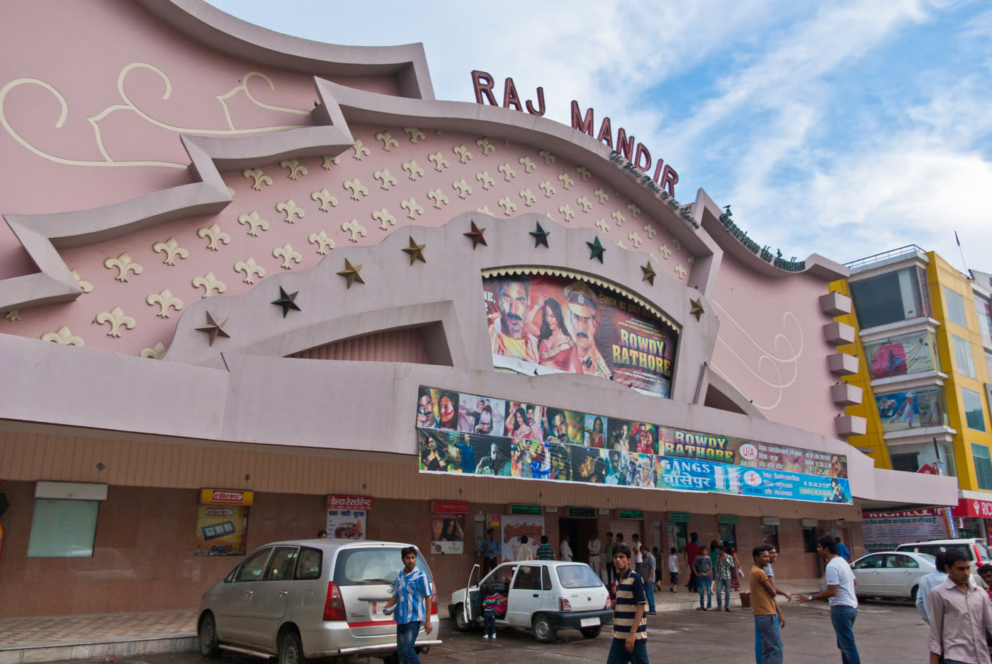 The pink art-deco facade of the Raj Mandir cinema in Jaipur, with painted film hoardings above the entrance and a crowd on the street outside.