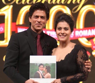 Shah Rukh Khan and Kajol standing together holding a commemorative book, in front of an illuminated backdrop reading "Celebrating 1000".